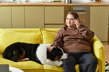 Down sendromlu genç bir kadın köpeğiyle birlikte parlak sarı bir kanepede eğleniyor..