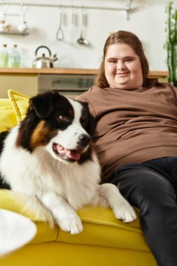 Down sendromlu genç bir kadın evinde köpeğiyle kanepede dinlenirken gülümsüyor..