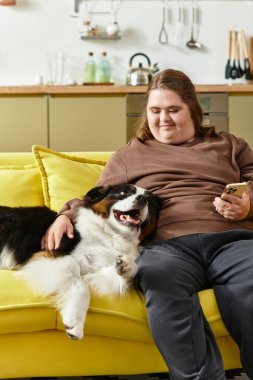 Gülümseyen genç bir kadın, parlak sarı bir kanepede rahatlar, köpeğiyle eğlenceli anlar geçirir..
