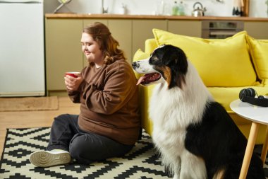 Genç bir kadın köpeğiyle yerde oturmuş sıcak bir içkinin tadını çıkarırken gülümsüyor..