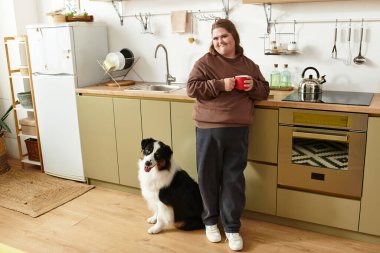 Down sendromlu genç bir kadın evde vakit geçirmekten zevk alıyor, elinde bir fincan çayla bir köpeğin yanında..