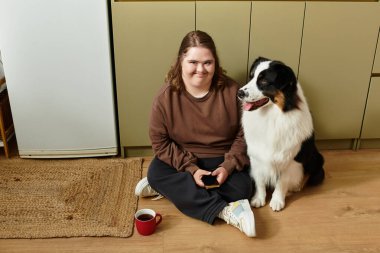 Genç bir kadın sıcak bir ev ortamında kürklü arkadaşının yanında gülümseyerek yerde dinleniyor..