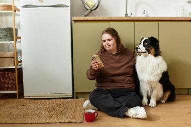 Neşeli genç bir kadın köpeğiyle yerde oturup sıcak ve davetkar bir şekilde telefonunu kontrol ediyor..