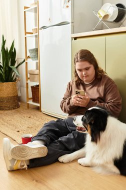 Down sendromlu genç bir kadın yerde oturuyor, köpeğiyle birlikte telefonuna odaklanmış..