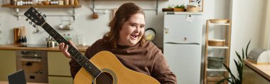 Down sendromlu genç bir kadın elinde gitarla evde müzik dinlerken gülümsüyor..