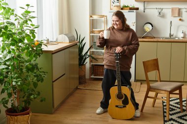 Down sendromlu genç bir kadın elinde bardak, yanında gitar tutarken gülümsüyor..