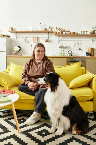 Down sendromlu genç bir kadın, yanındaki köpeğine nazikçe gülümseyen sarı bir koltukta oturuyor..