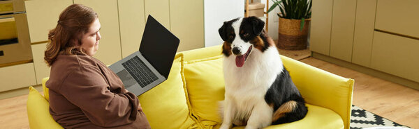 Молодая женщина с синдромом Дауна сидит на жёлтом диване, сосредоточенная на ноутбуке со своей собакой.