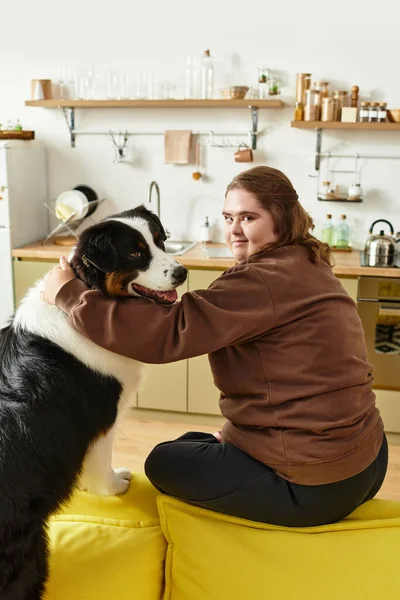 Genç bir kadın, sıcak bir mutfakta köpeğiyle mutlu bir şekilde, parlak bir kanepede oturuyor..