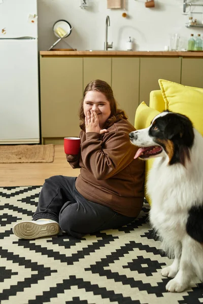Down sendromlu mutlu genç bir kadın köpeği ve bir kupayla yerde dinleniyor..