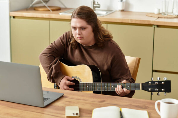 Молодая женщина с синдромом Дауна играет на гитаре за своим кухонным столом, сосредоточенная на ноутбуке.