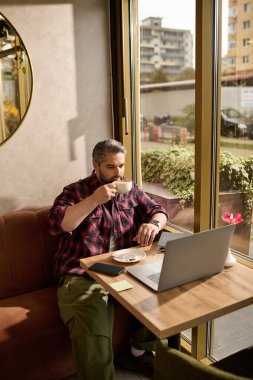 Parlak bir kafede dizüstü bilgisayarla çalışırken şık giyinmiş olgun bir adam kahvesini yudumluyor..