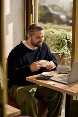 Şık bir kafede yakışıklı bir adam bilgisayarına ve notlarına odaklanırken kahvesinin tadını çıkarıyor..