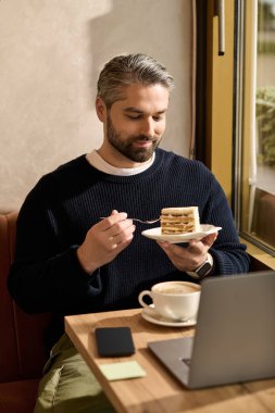 Yakışıklı bir adam sıcak bir kafede laptopuyla uğraşırken bir dilim kek ve kahvenin tadını çıkarıyor..