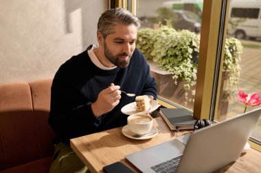 Yakışıklı adam kek ve kahvenin tadını çıkarıyor, sıcak, davetkar bir kafe ortamında laptopuna odaklanmış..