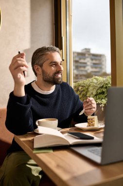 Yakışıklı, olgun bir adam, rahat bir kafede işine odaklanırken kek ve kahvenin tadını çıkarıyor..