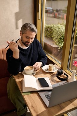 Bir masada oturmuş, kahvesini yudumluyor ve sıcak bir kafe ortamında tatlısını yiyor..