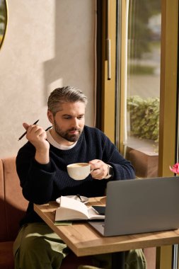 Şık giyinmiş olgun bir adam kahvesini yudumlar ve sakin bir kafede dizüstü bilgisayarına odaklanır..