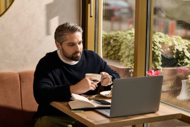 Yakışıklı, olgun bir adam kahvesini yudumlar ve büyüleyici bir kafede dizüstü bilgisayarıyla çalışır..