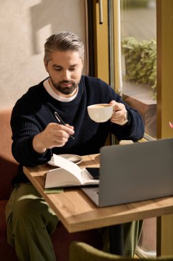 Yakışıklı, olgun bir adam bir masada oturur, kahvenin tadını çıkarırken aynı zamanda bir not defterine notlar yazar..