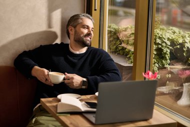 Olgun ve yakışıklı bir adam, pencerenin yanında laptopuyla çalışırken bir fincan kahveyle rahatlar..