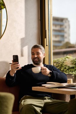 Yakışıklı, olgun bir adam, şık giyinmiş telefonuna gülümserken kahvesini yudumluyor..