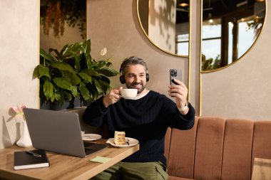 Havalı giyinmiş yakışıklı bir adam elinde kahve fincanı tutarken bir yandan gülümsüyor, bir yandan da modaya uygun bir kafede görüntülü sohbet yapıyor..
