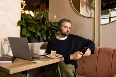 Yakışıklı olgun bir adam şık bir kafede oturur, telefonuna odaklanarak kahve ve tatlıdan zevk alır..