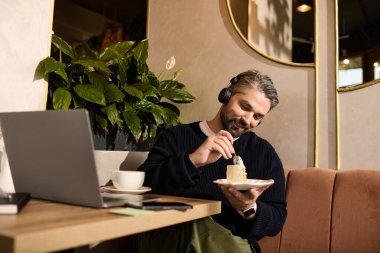 Olgun bir adam, zengin bitkilerle çevrili zarif bir kafede müzik dinlerken pastanın tadını çıkarır..