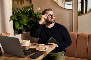 Tarz sahibi, kendine güvenen bir adam dizüstü bilgisayarında kek ve kahvenin tadını çıkarırken gülümser..