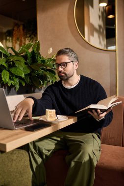 Moda giyinmiş olgun bir adam laptopla meşgul olurken tatlı yiyor ve kafede kitap okuyor..