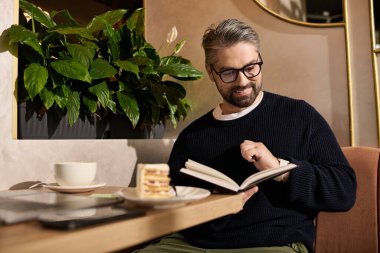 Olgun, yakışıklı bir adam şık bir kafe atmosferinde tatlı yerken kitap okur..