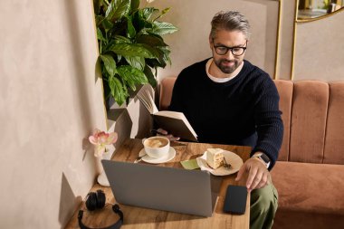 Yakışıklı adam kitap okuyor, kahvesini yudumluyor ve şık bir kafe ortamında tatlının tadını çıkarıyor..