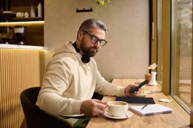 Şık giyinmiş olgun bir adam ahşap bir masada oturur ve telefonunu kullanarak kahvesini yudumlar..