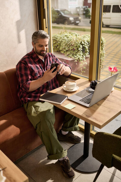 Handsome mature man relaxes in a cozy cafe, savoring coffee while using his smartphone.