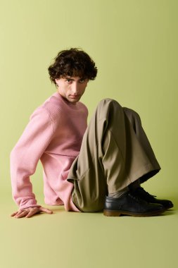 Pembe kazaklı ve rahat pantolonlu, kendine güvenen bir adam yerde oturuyor, cazibesi ve tarzı var..