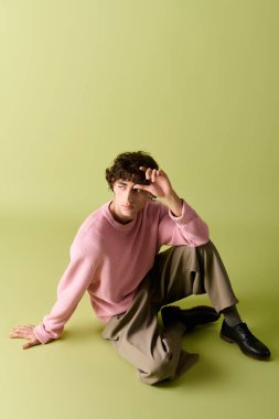 Modaya uygun bir adam yerde poz veriyor, bir eline yaslanıyor, pembe bir kazak giyiyor..