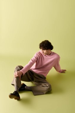 Pembe kazak ve şık pantolon giymiş yakışıklı bir adam zarif bir şekilde yeşil zeminde oturuyor..