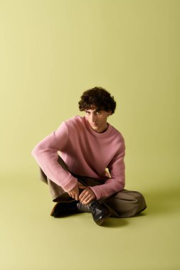 Pembe kazaklı ve şık pantolonlu yakışıklı bir adam yeşil bir zemine karşı düşünceli bir şekilde poz verir..