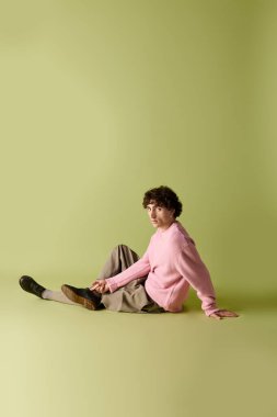 Moda kıyafetleri içinde yakışıklı bir adam açık yeşil bir arkaplanda sakince poz veriyor, zarafet saçıyor..