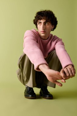 Pembe kazaklı ve şık pantolonlu yakışıklı bir adam yumuşak yeşil bir zeminde düşünceli bir şekilde çömeliyor..