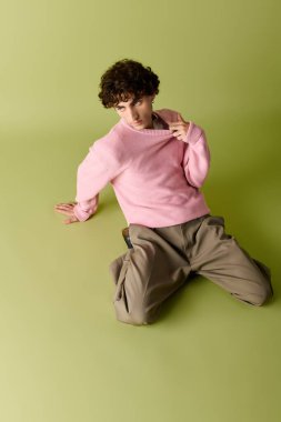 Şık bir adam pembe bir süveter ve rahat bir pantolonla yerde oturuyor, kendinden emin..