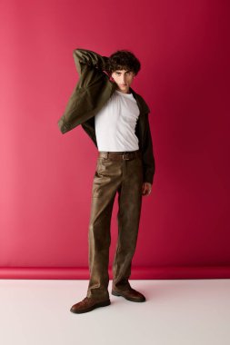 Şık bir adam kendine güvenen bir duruş sergiliyor. Pembe bir arka plan karşısında modaya uygun kıyafetler giyiyor..