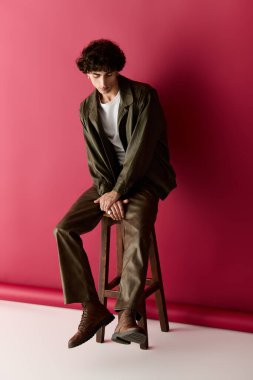 Yakışıklı adam bir taburede otururken modern giysiler sergiliyor, zarafet ve cazibe yayıyor..