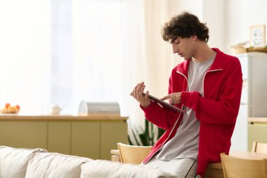 Moda kıyafetleri içinde yakışıklı bir adam güneşli bir odada otururken tabletini okuyor..