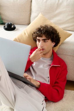 Moda kıyafetleri içinde odaklanmış genç adam dizüstü bilgisayarı ve aperatifleri kullanarak yerde uzanıyor..