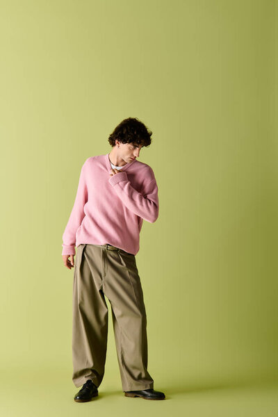 Красивый мужчина в розовом свитере и свободных брюках уверенно стоит на фоне.