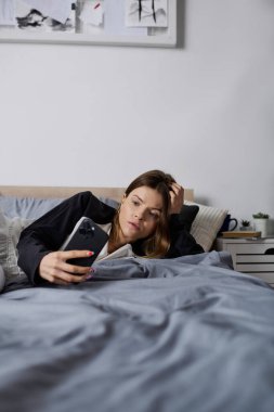 Genç bir kadın, sakin bir günün tadını çıkarırken telefonunu kontrol ederek yatakta dinleniyor..