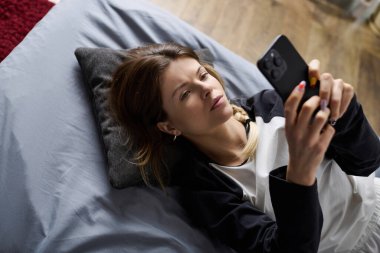 Genç bir kadın, sakin bir günü yatağında, telefonunu modern bir yerde dinlenerek geçirir..