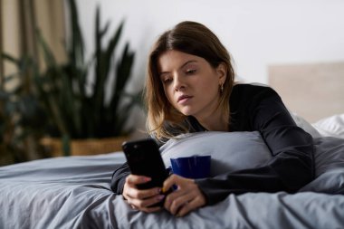 Genç bir kadın gününü yatakta geçiriyor, rahat bir dairede kahvesini yudumlarken telefonunu kaydırıyor..